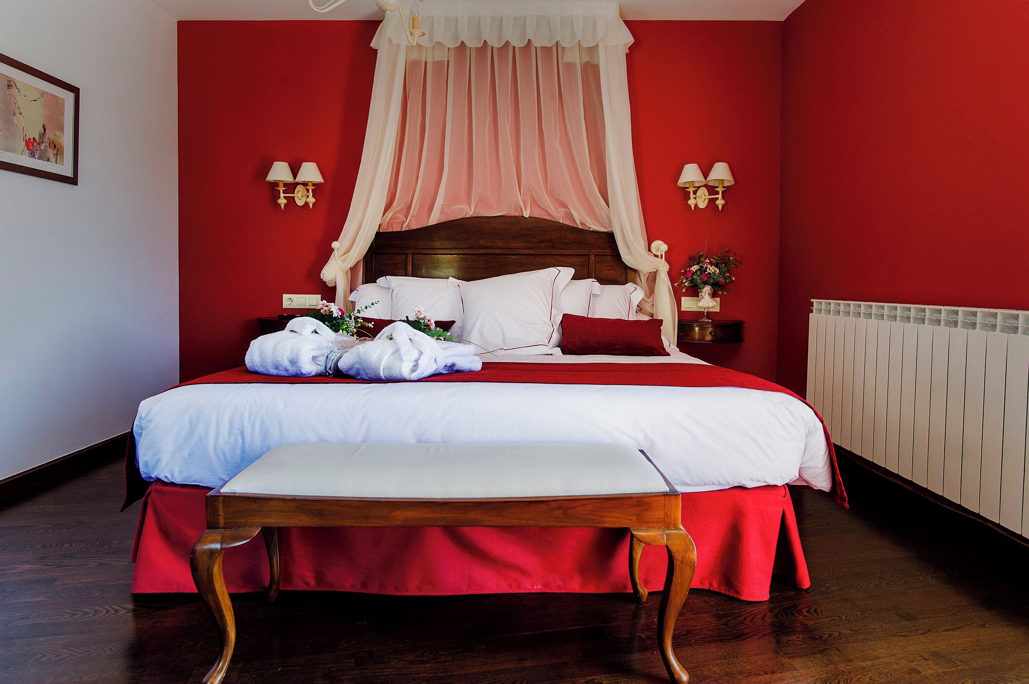 A neatly made bed with white linens and a red bedspread, a wooden bench at the foot of the bed, and a red wall with a white radiator.