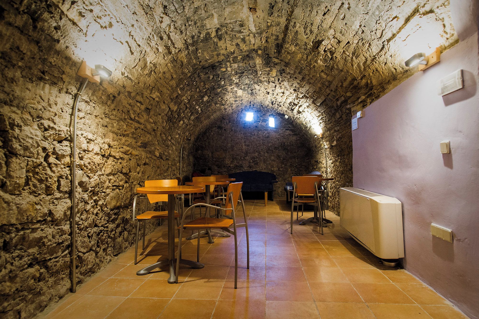 A stone-walled interior with tables and chairs, a radiator, and a small table lamp.