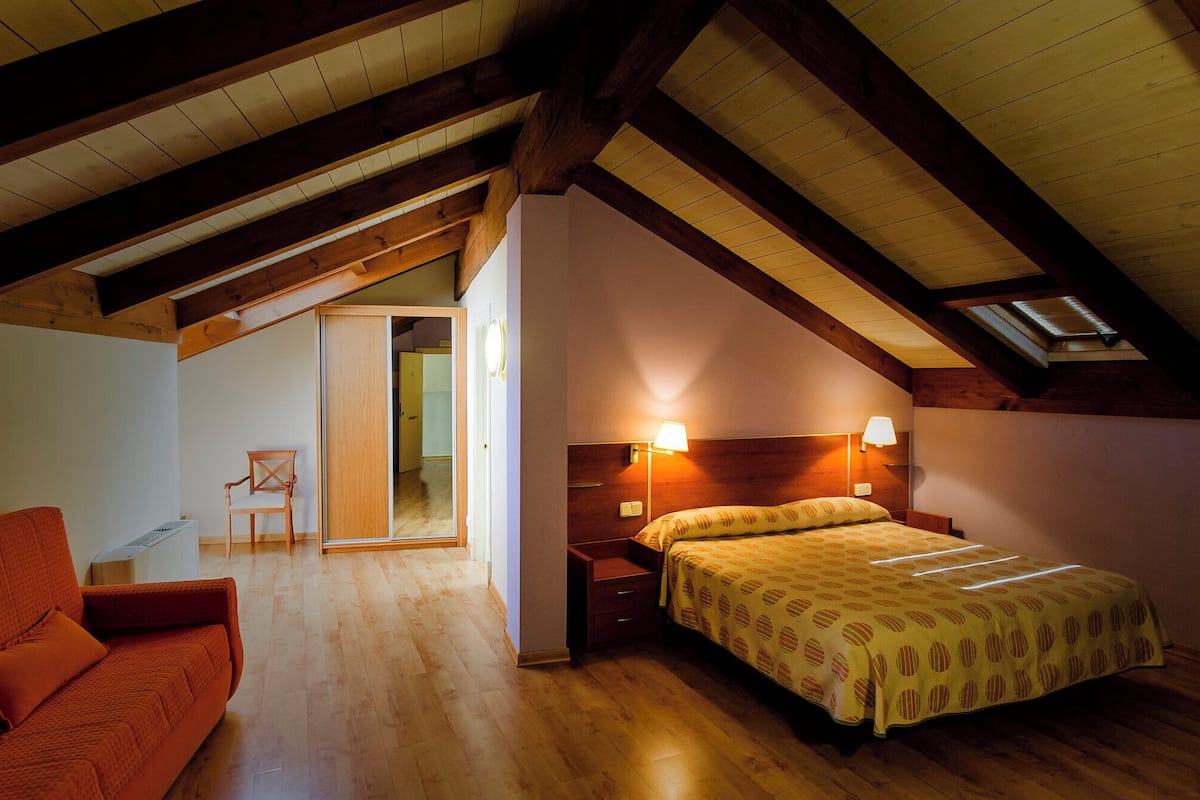 Une chambre avec un plafond en bois, un lit avec une literie à motifs, une table de chevet et une chaise.