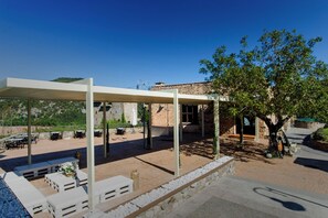 An outdoor patio with white tables and benches, a covered seating area, and a stone building in the background. - Ligueerre Resort (Abizanda)