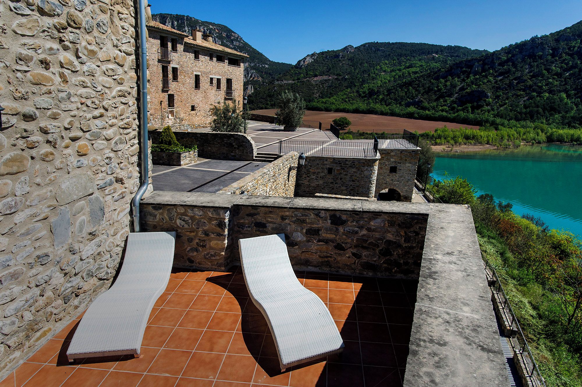 A stone building with a terrace featuring two lounge chairs and a view of a lake and mountains.