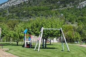 A playground with a swing set and climbing equipment, surrounded by grass and trees, with a mountainous backdrop. - Ligueerre Resort (Abizanda)