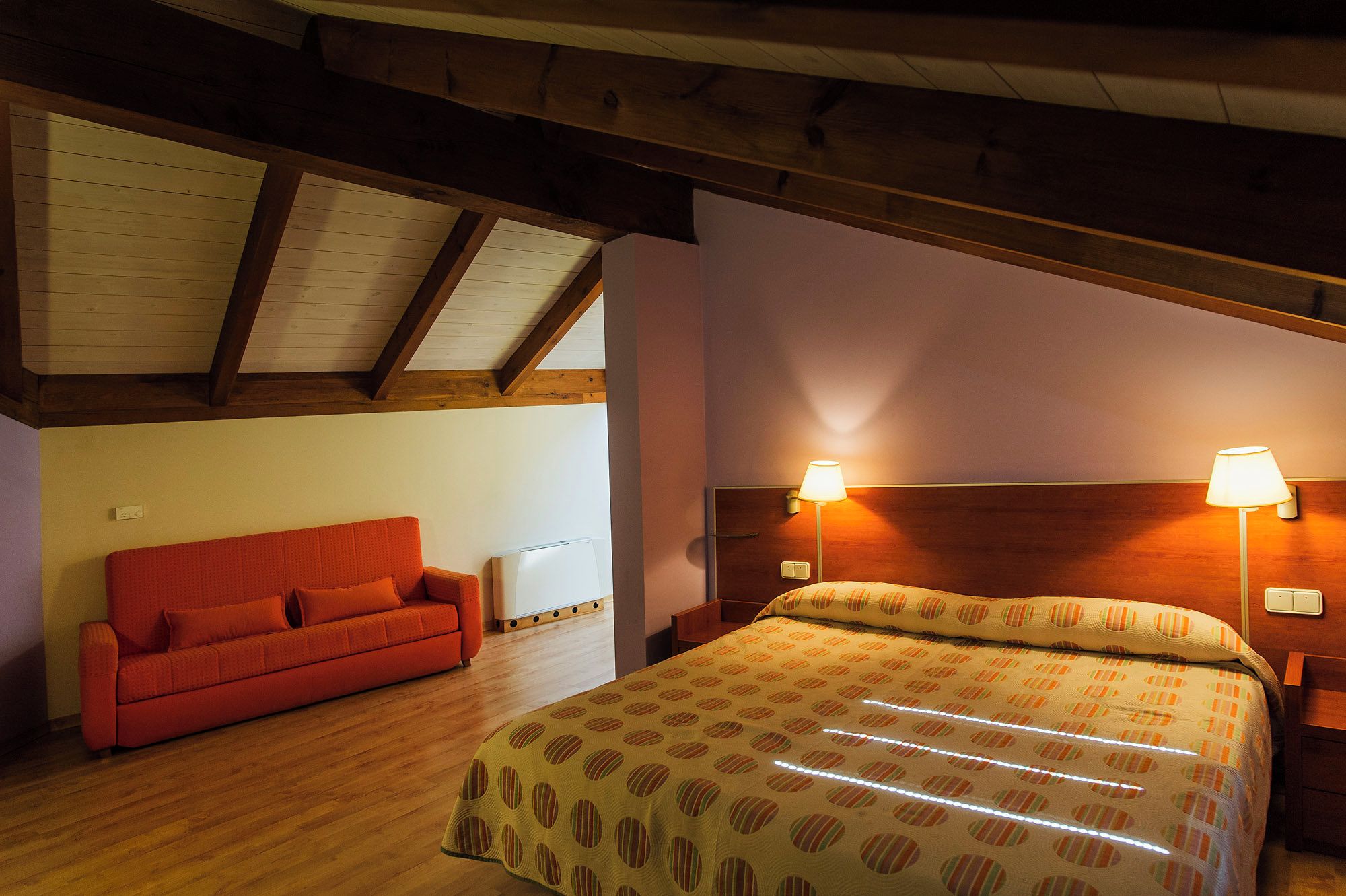A bedroom with a wooden ceiling, a red sofa, a bed with a patterned bedspread, and two bedside lamps.