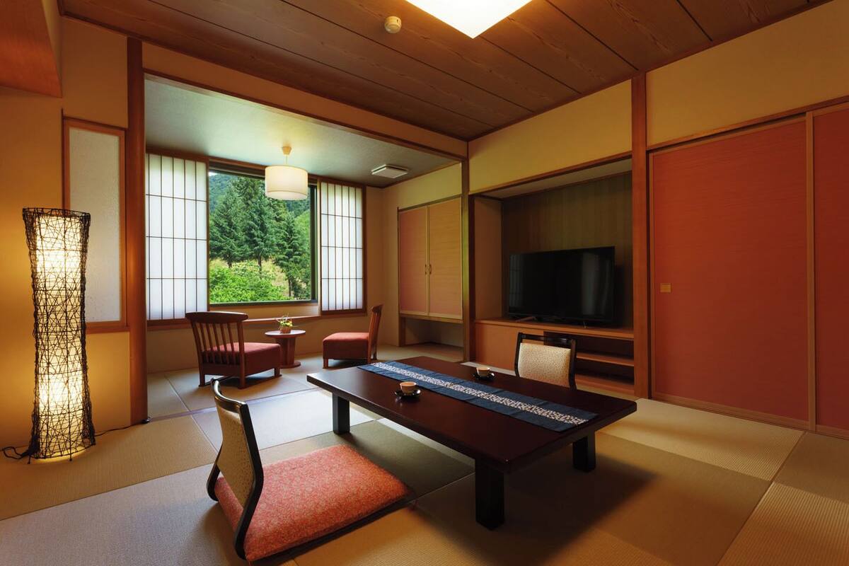A traditional Japanese-style room with tatami flooring, a low table, chairs, a television, and a large window with a view of greenery.