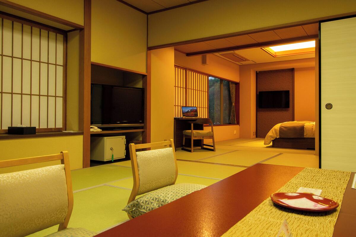 A traditional Japanese room with tatami flooring, a low table, chairs, a television, and a bed.