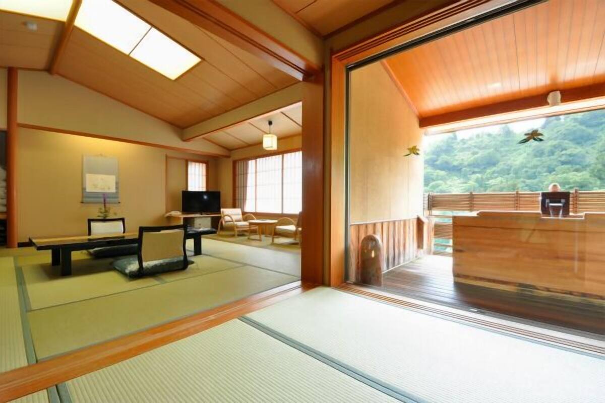 A traditional Japanese room with tatami flooring, a low table, a television, and a view of a forested area.
