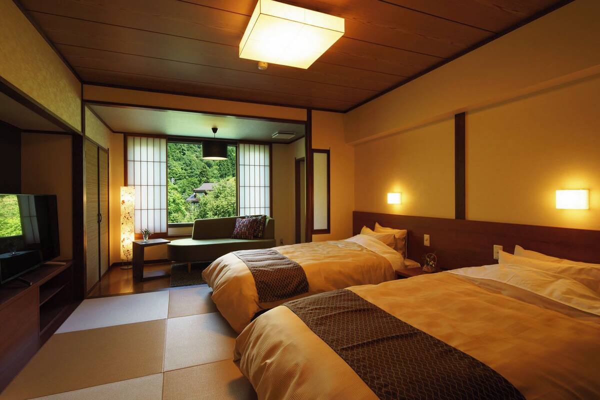 A hotel room with two beds, a TV, a desk with a chair, and a view of greenery outside.