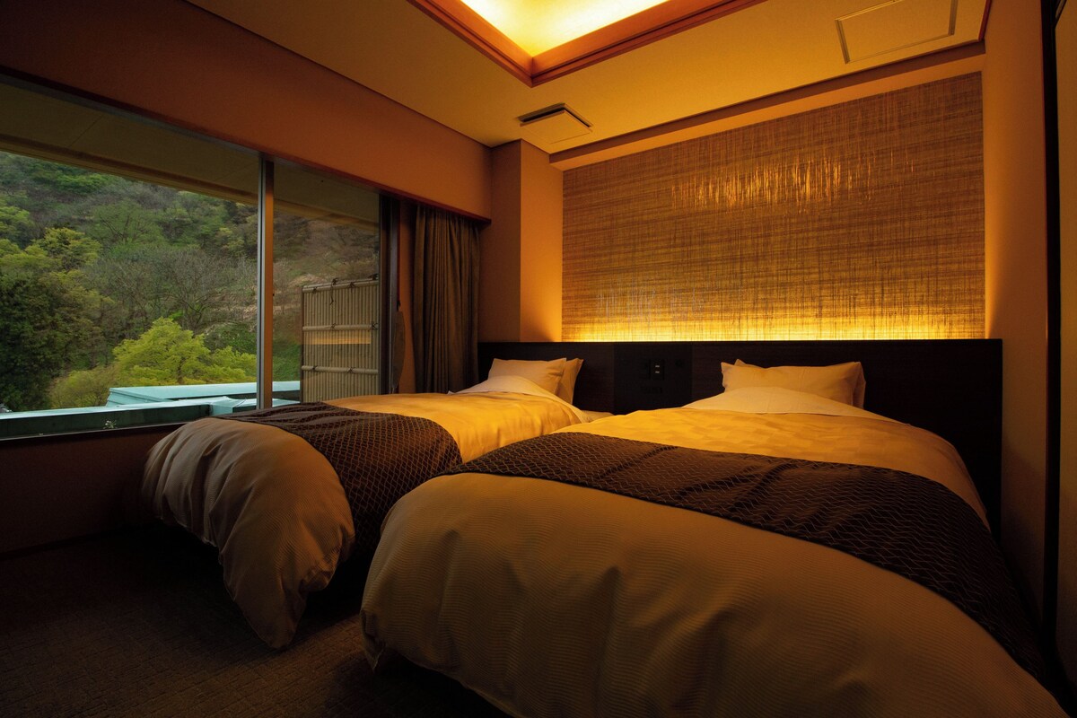 A hotel room with two beds, a large window with a view of greenery, and a wall-mounted light fixture.