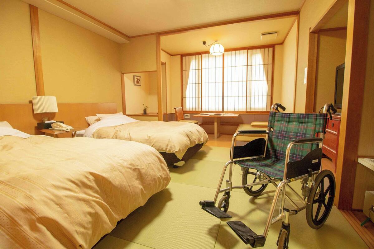 A hotel room with two beds, a wheelchair, a bedside table, a lamp, and a window with curtains.