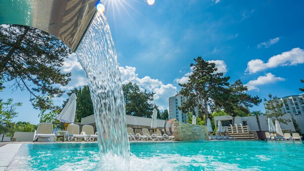 En swimmingpool med et vandfald, omgivet af liggestole og parasoller, under en klar blå himmel.