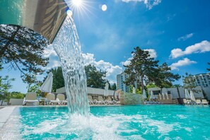 A swimming pool with a waterfall feature, surrounded by lounge chairs and umbrellas, under a clear blue sky. - Mera Resort Venus (Venus)