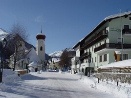 Calle nevada con una torre de iglesia, Hotel Pension Kirchplatz y edificios tradicionales.