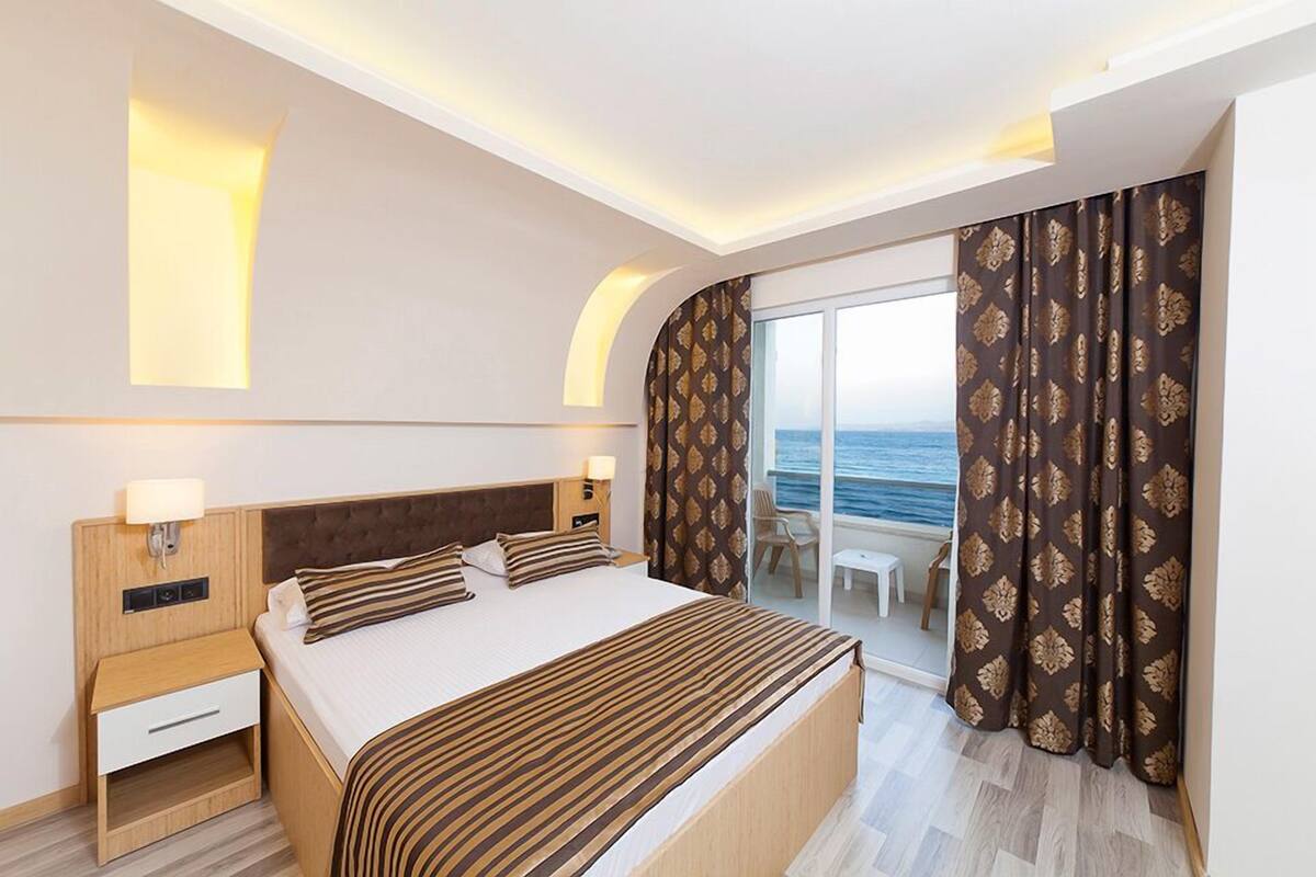 A modern hotel room with a large bed, bedside tables, a nightstand, and a view of the sea through a sliding glass door.