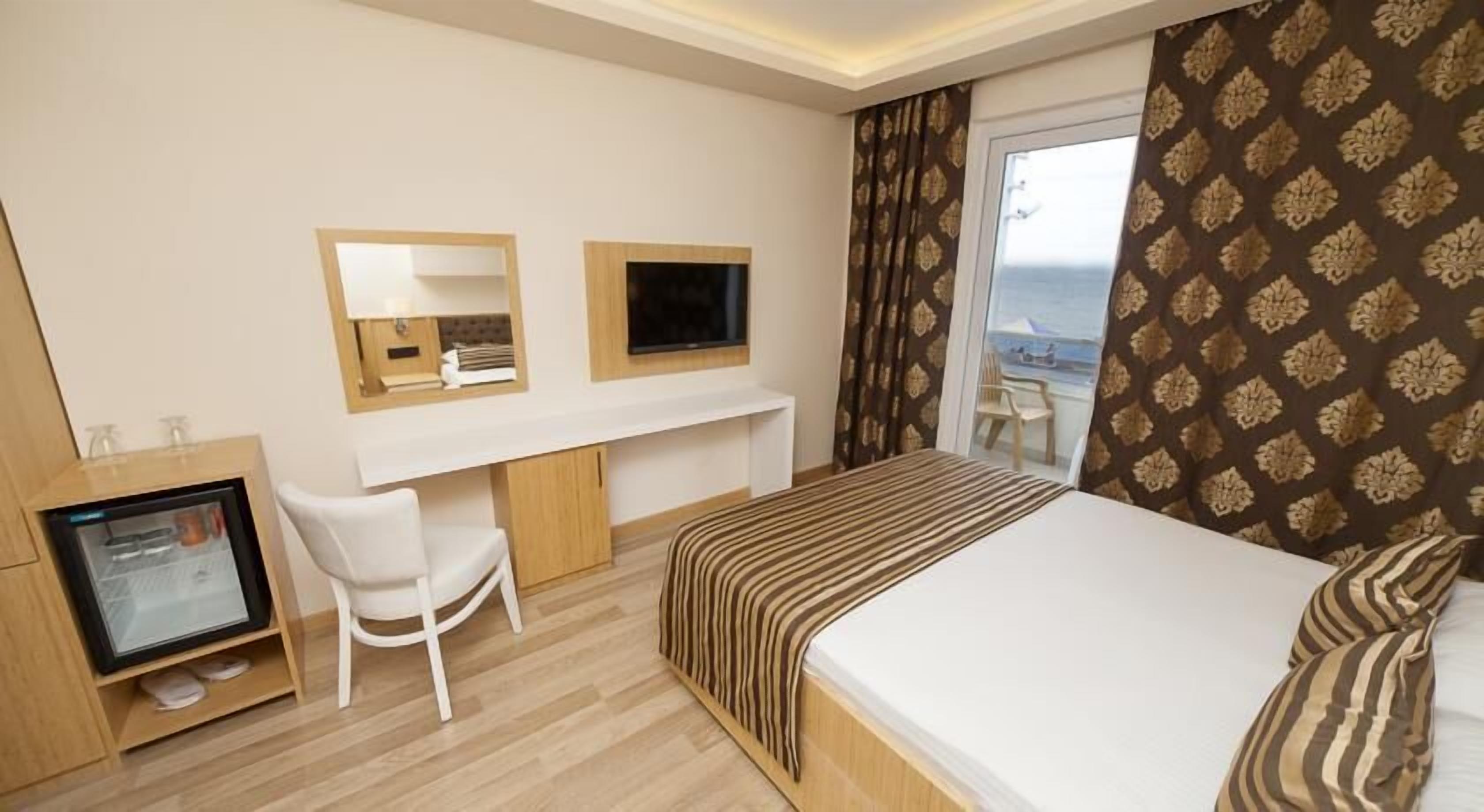 A hotel room with a bed, a desk with a computer, a chair, a TV, and a window with a view of the sea.