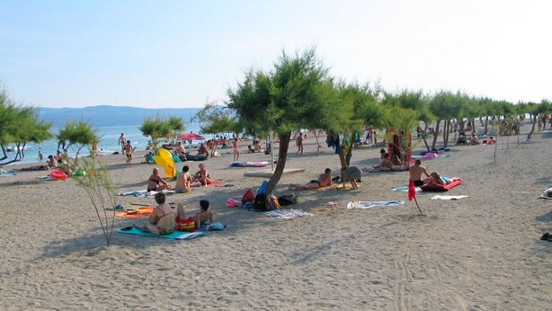A beach with people sunbathing and swimming, trees providing shade, and a clear sky. - Mladen (Omis)