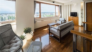 A modern living room with a sofa, armchair, and coffee table. There is a television on a wooden cabinet and a large window with a view of the city. - Hokkaido Hotel (Obihiro)