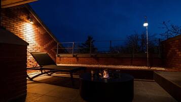 An outdoor patio area with a brick wall, a lounge chair, a round table with a fire feature, and a street lamp.