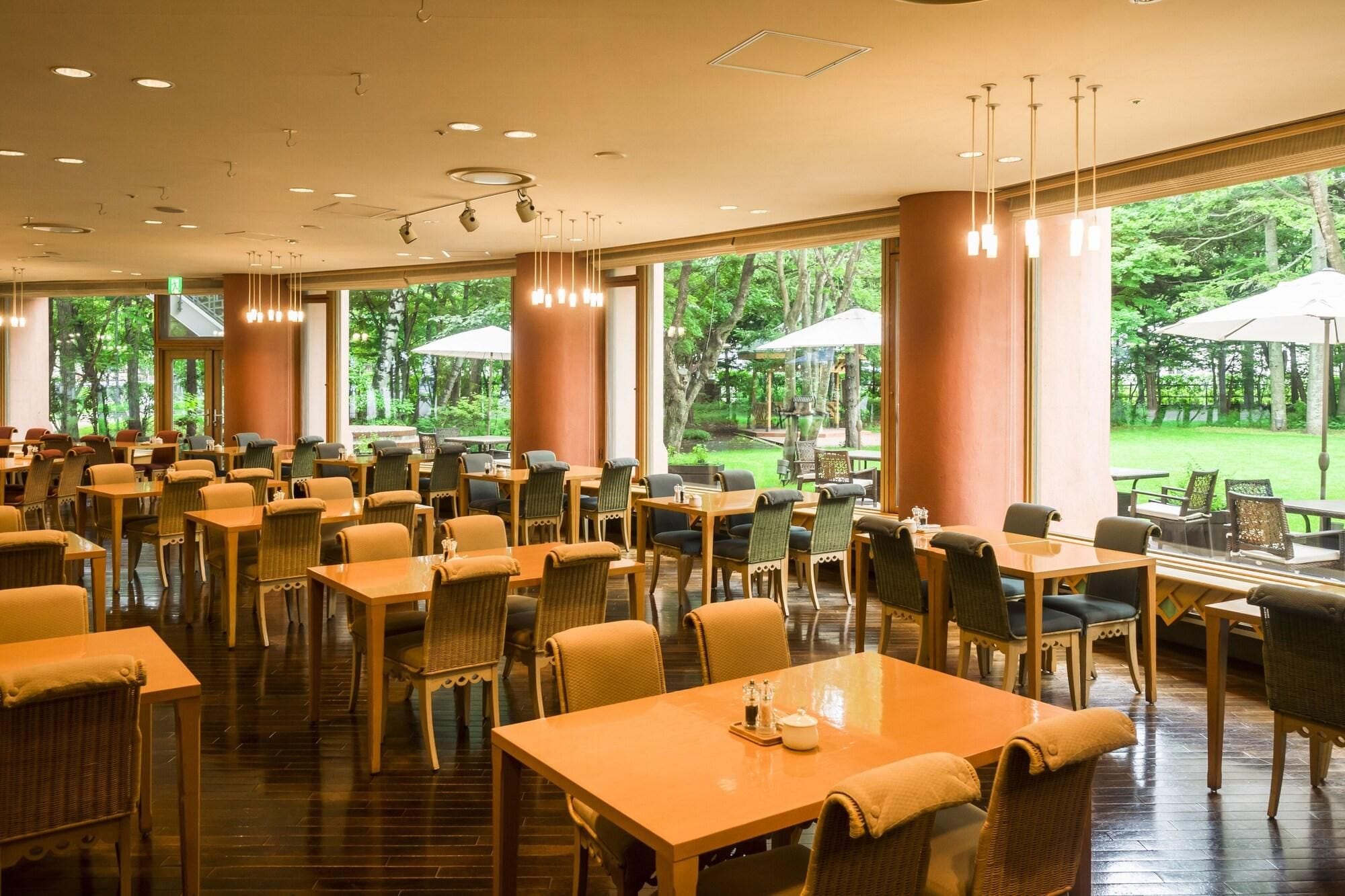 A spacious indoor dining area with multiple tables and chairs, large windows offering a view of the outdoors, and ceiling lights.