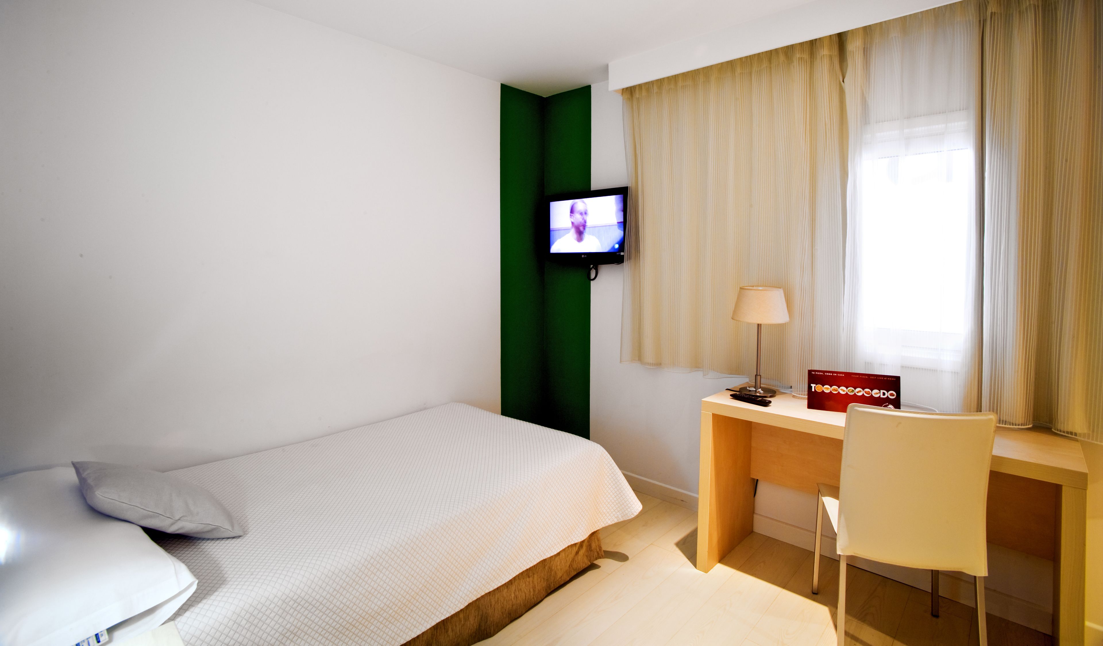 A hotel room with a bed, a desk with a chair, a television mounted on the wall, and a window with curtains.