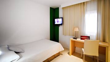 A hotel room with a bed, a desk with a chair, a television mounted on the wall, and a window with curtains.