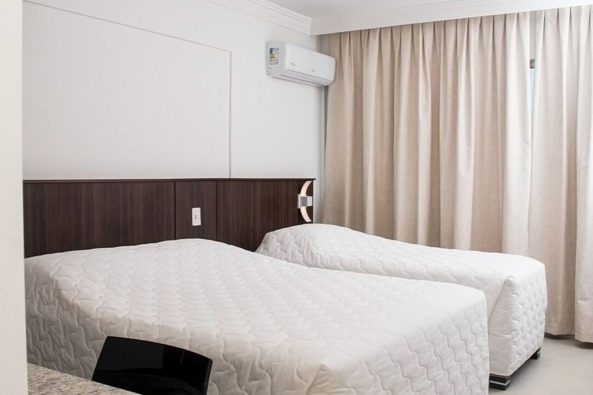 A hotel room with two beds, a dark wood headboard, a white air conditioning unit, and beige curtains.