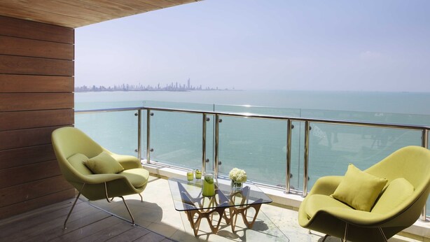 A balcony with two green chairs, a glass table, and a view of the sea and a distant city skyline.