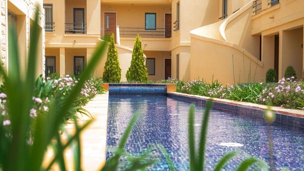 Une piscine dont le bord est recouvert de carreaux, entourée de verdure et de fleurs, avec un immeuble à plusieurs étages en arrière-plan.