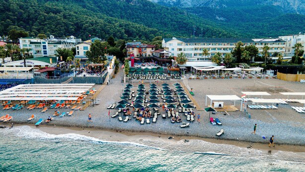 A beach with rows of sun loungers and umbrellas, a hotel in the background, and a mountainous landscape. - Aleria Belport Beach Hotel - All inclusive (Kemer)