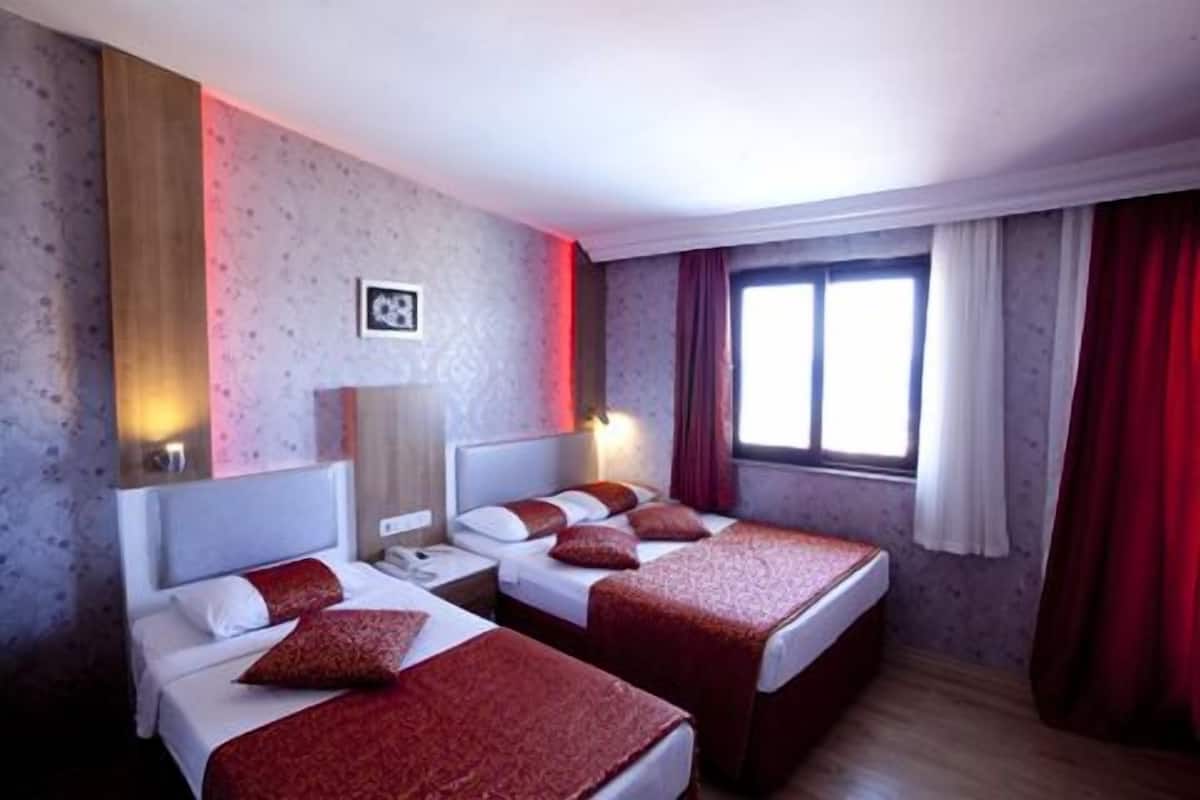 A hotel room with two beds, a nightstand, a lamp, a window with curtains, and a wall-mounted picture.
