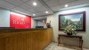 Un área de recepción con un letrero rojo de 'RAMADA', un mostrador de madera, piso de baldosas y un cuadro enmarcado con una vista urbana.