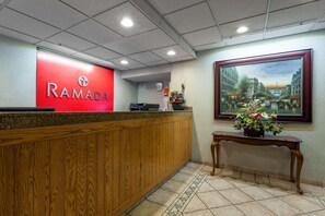 A reception area with a red 'RAMADA' sign, a wooden reception desk, a tiled floor, and a framed painting of a cityscape. - Ramada by Wyndham Seatac Airport North (Tukwila)