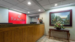 A reception area with a red 'RAMADA' sign, a wooden reception desk, a tiled floor, and a framed painting of a cityscape.