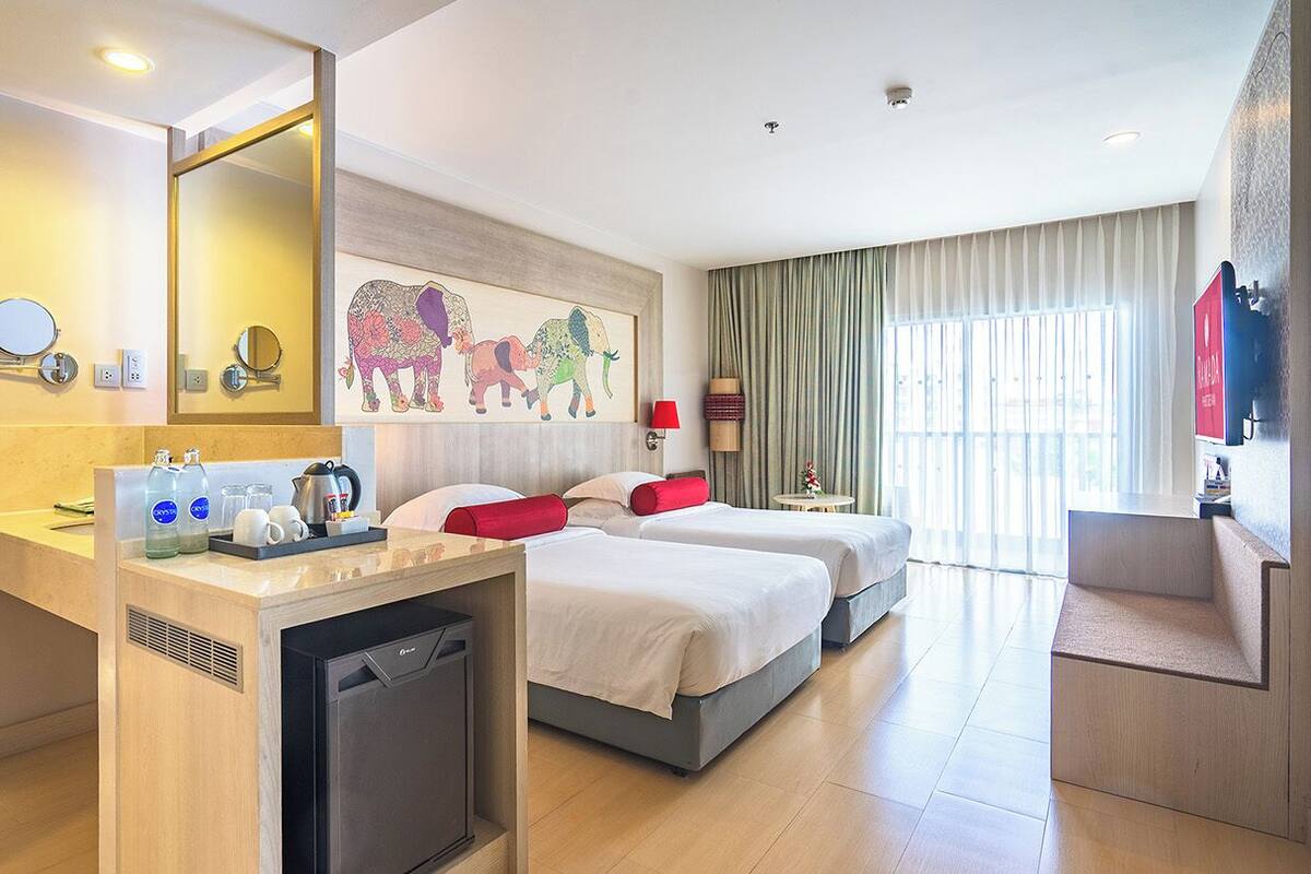 Een hotelkamer met een bed, nachtkastjes, een bureau met een koelkast, een spiegel en een schilderij van olifanten aan de muur.