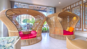 Un intérieur moderne avec deux fauteuils en osier aux formes arrondies, dotés de coussins rouges, un canapé en osier et une vue sur l’extérieur grâce à de grandes fenêtres.