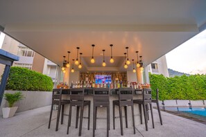 Zona de bar al aire libre con sillas de mimbre, piscina y barra repleta de botellas.