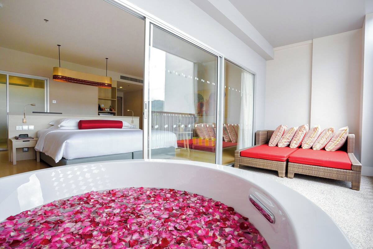 Een moderne hotelkamer met een groot, wit bad vol rozenblaadjes, een bed met witte lakens en een rode kussen, een rieten bank met rode kussens, en een grote schuifglazen deur naar een balkon.