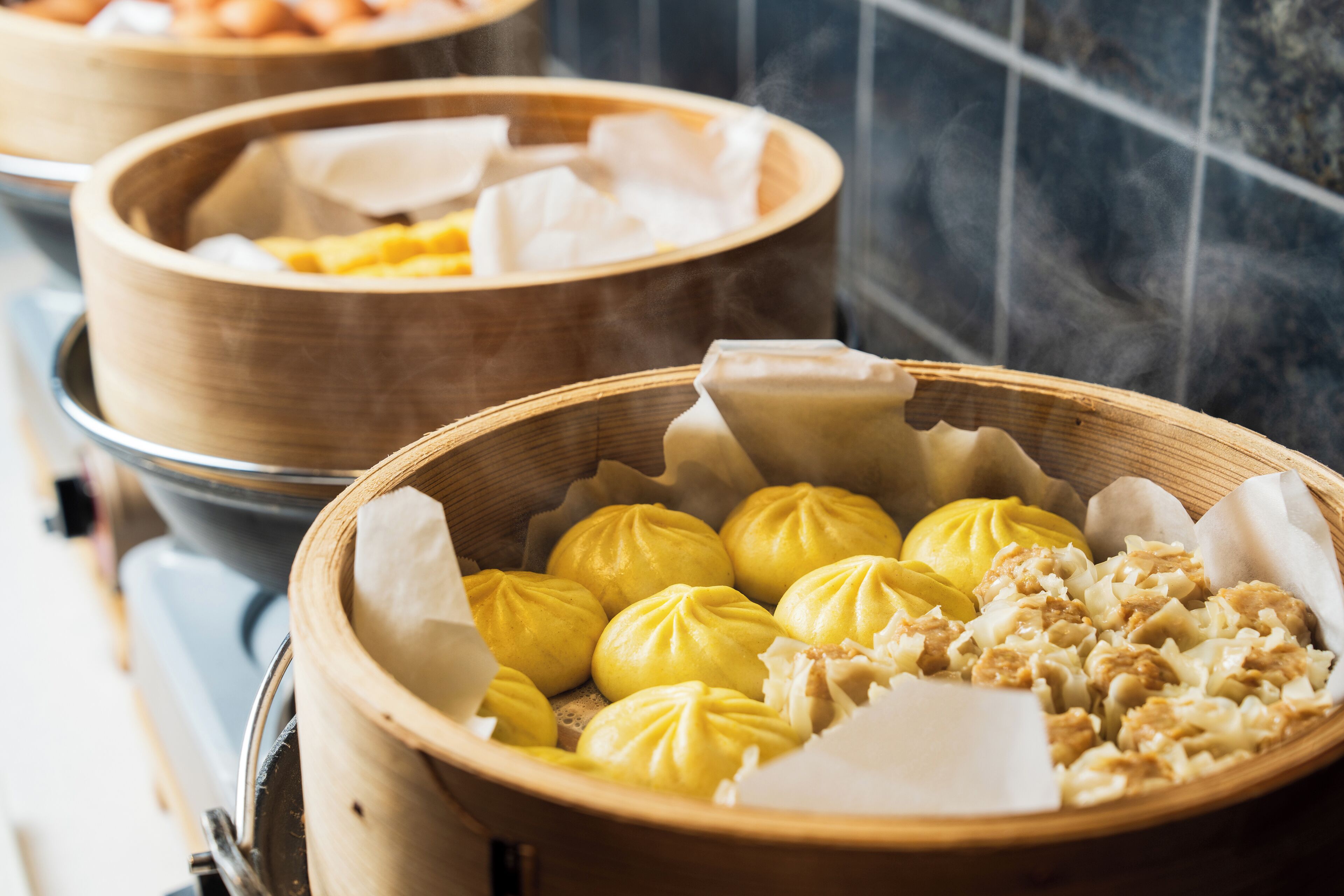 A close-up of a bamboo steamer basket filled with yellow steamed buns and a wooden tray with white dumplings.