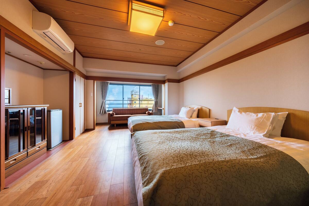A hotel room with a wooden ceiling, a large bed, a smaller bed, a desk, and a window with a view of the sky.