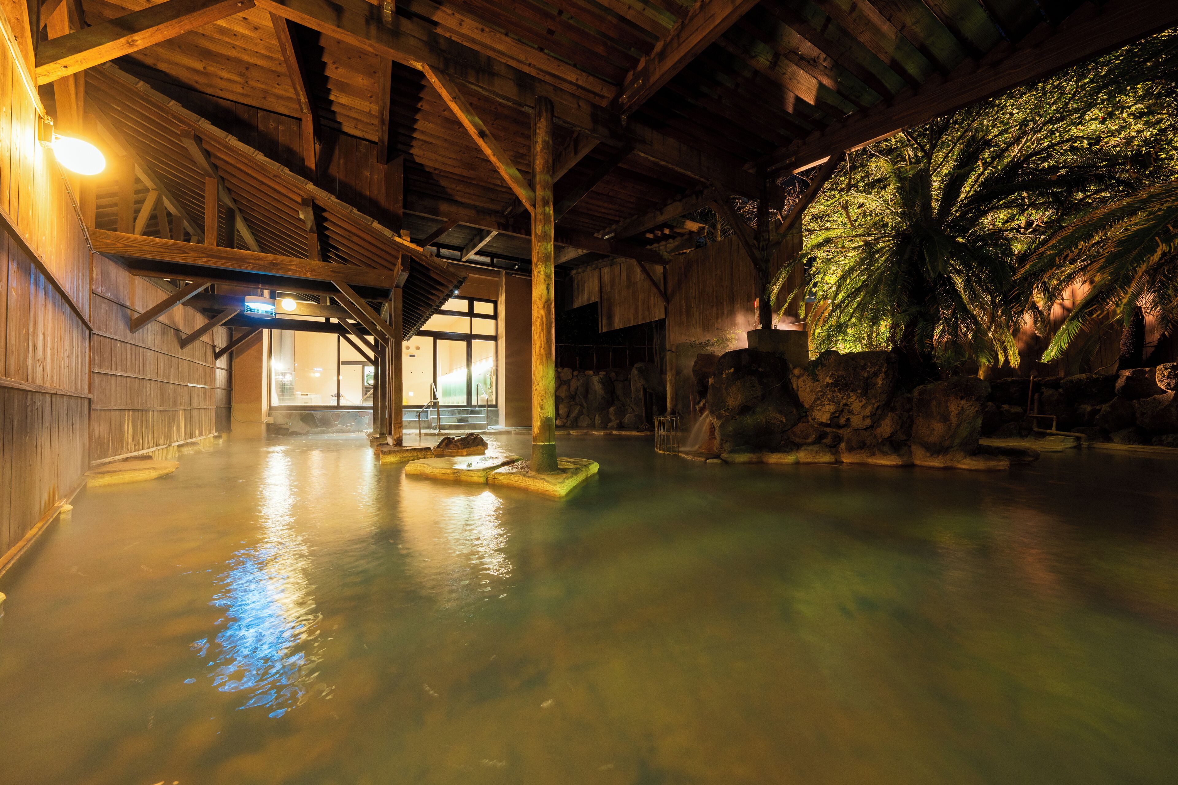 A traditional Japanese onsen with wooden architecture, a hot spring pool, and a large tree providing shade.