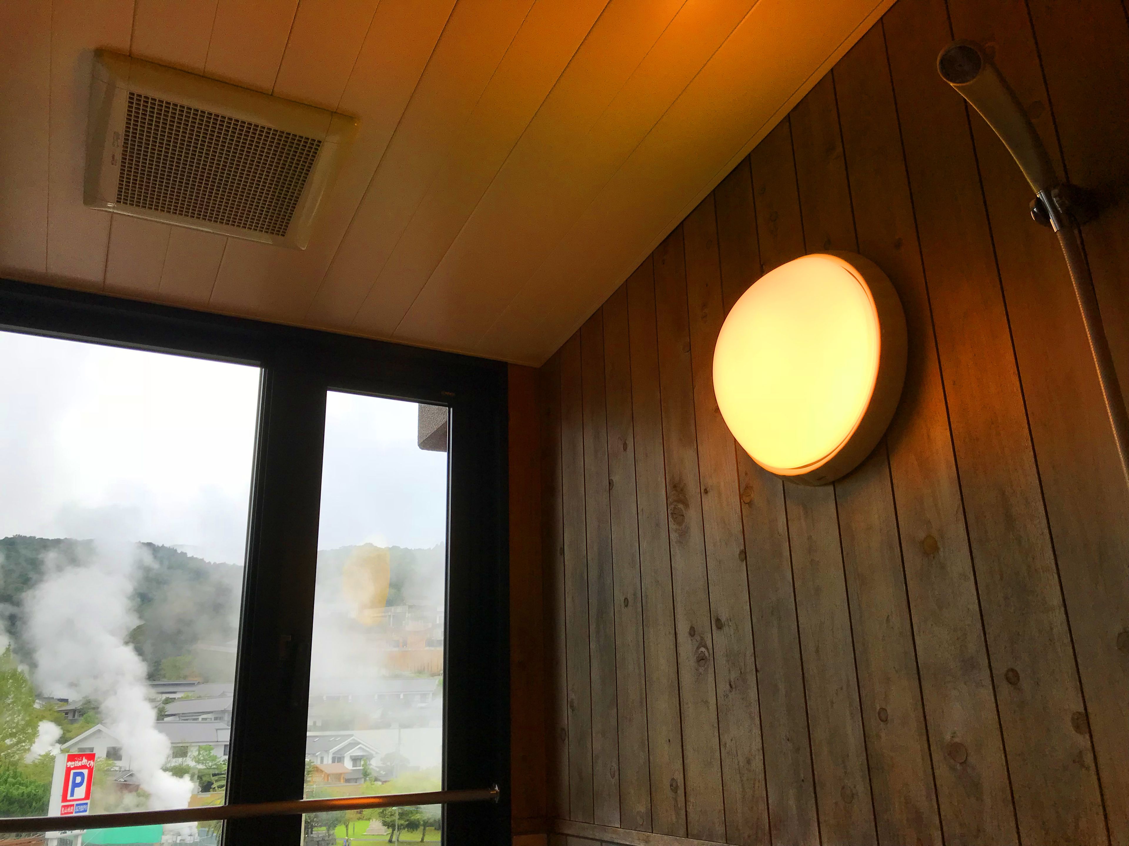 A room with wooden walls, a round light fixture, a window with a view of buildings and smoke, and a ceiling vent.