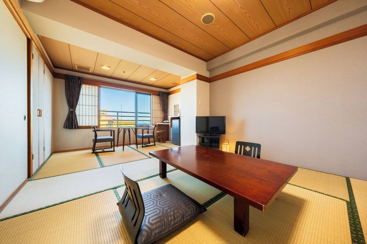 A traditional Japanese room with tatami flooring, a wooden ceiling, a dining table, a TV, and a view of the outside.