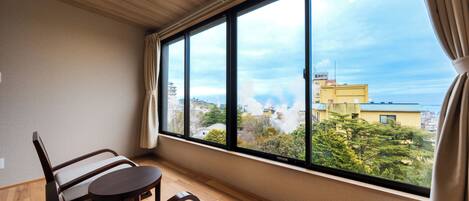 A room with a large window, a wooden ceiling, a sofa, a small table, and a view of buildings and trees.