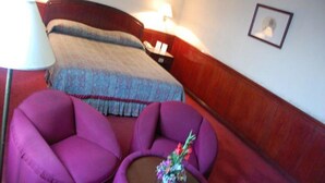 A hotel room with a bed, two purple armchairs, a small table with a vase, and a nightstand with a lamp.