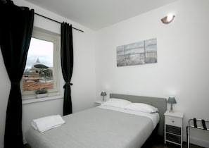 A neatly made bed with white linens, a bedside table with a lamp, and a view of a cityscape through a window with black curtains. - Candia Green House B&B (Rome)