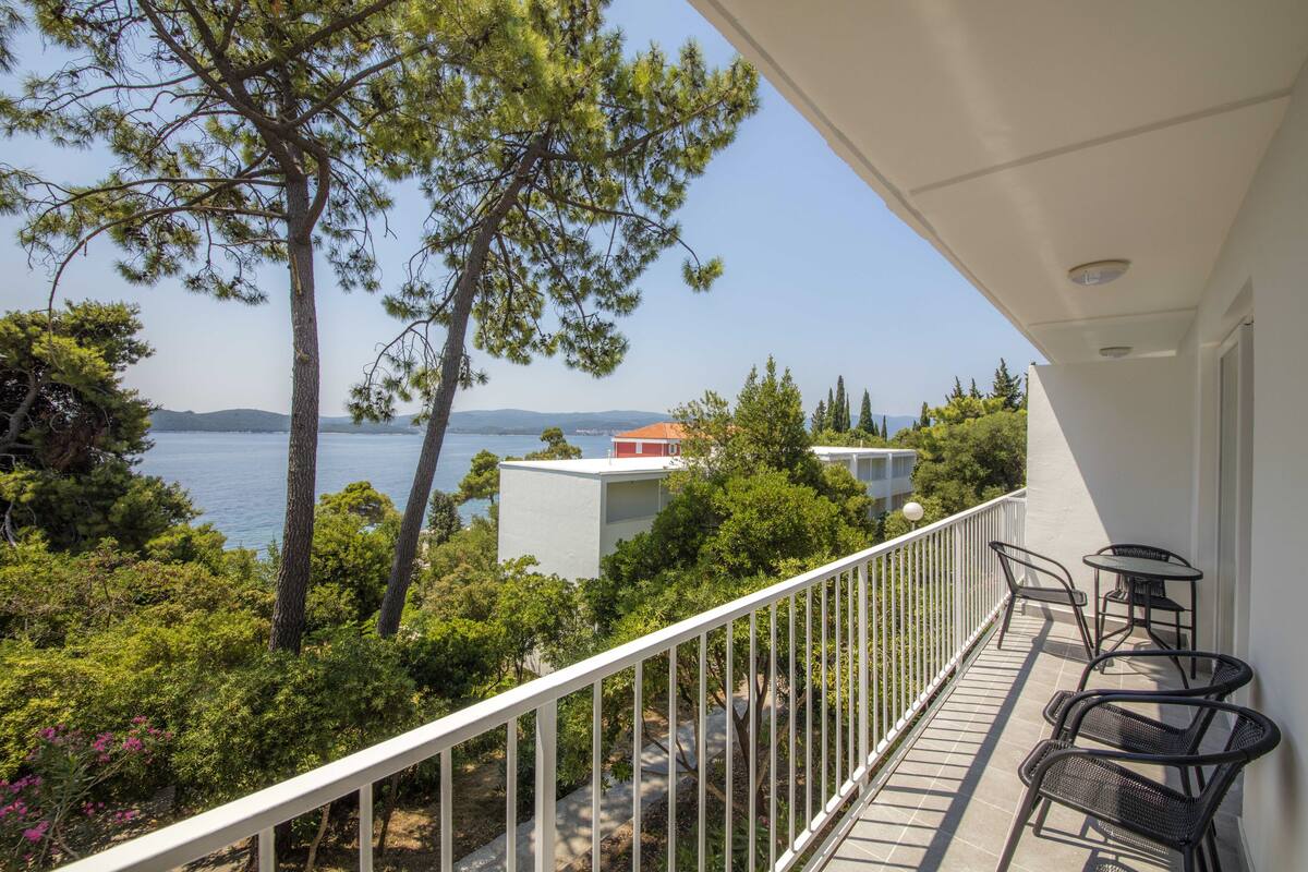 A balcony with a white railing, overlooking a sea view, surrounded by greenery and trees.