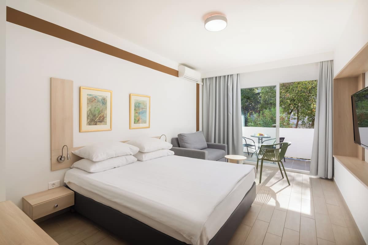A modern hotel room with a large bed, a desk, a chair, and a balcony with a view of trees.