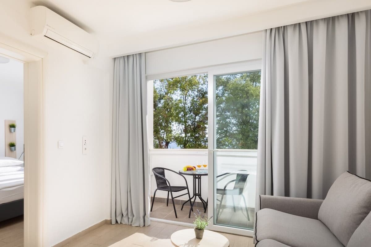 A modern hotel room with a bed, a sofa, a dining area with chairs, and a large window with curtains.