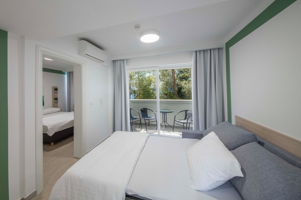 A hotel room with a bed, a balcony with chairs, and a view of trees.