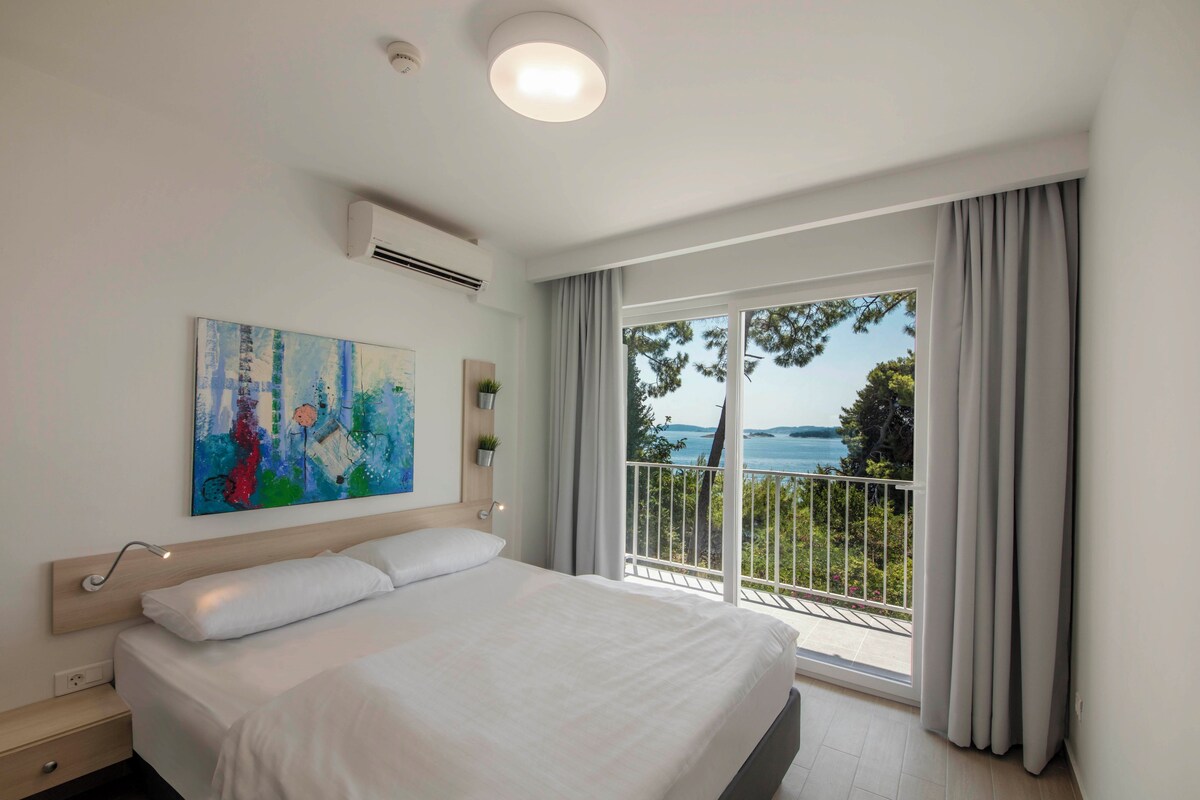 A bedroom with a bed, a painting, an air conditioning unit, and a balcony with a view.