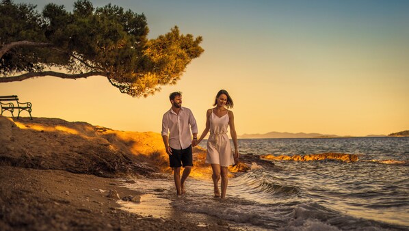 A couple walking along a rocky beach at sunset, holding hands, with a bench and a tree in the background. - Bellevue Maradiso Casa by Aminess (Orebic)